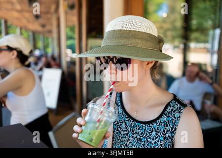 Junge Frau mit Sonnenhut und Sonnenbrille, die an einem Sommertag auf einer Außenterrasse eines Cafés durch einen Strohhalm eisgekühlten Matcha-Tee schlürft. Stockfoto