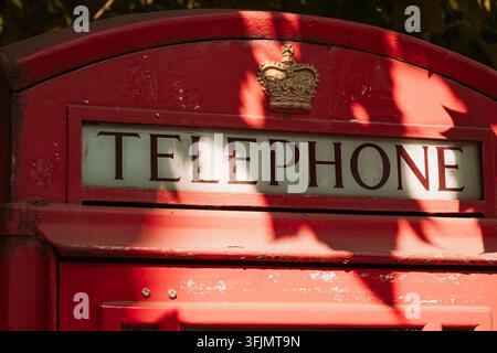 Detaillierte Nahaufnahme des oberen Rahmens eines traditionellen roten Telefonkastens, der das Wort TELEPHONE unter einer goldenen Krone mit Blattschatten anzeigt Stockfoto