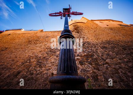 Direkte Blickwinkelaufnahme, die direkt nach oben zu einer Straßenlaterne schaut, die mit einem roten Fahrrad geschmückt ist, eingerahmt von einer alten Steinmauer unter einem klaren blauen Himmel. Stockfoto