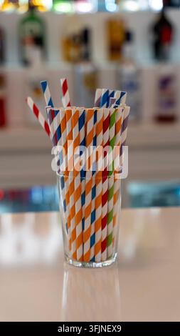 Bunte gestreifte Papier-Trinkhalme, die in einem Glas auf einer Theke angeordnet sind, mit verschwommenen Flaschen im Hintergrund. Stockfoto