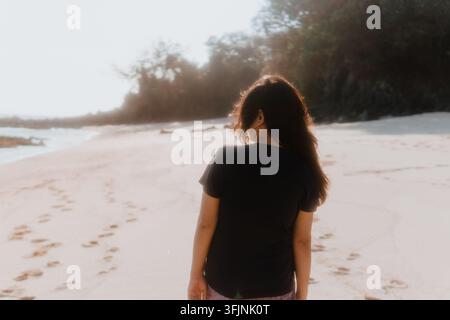 Rückansicht einer jungen Frau mit langen Haaren, die an einem sandigen tropischen Strand an einem hellen Tag steht. Stockfoto