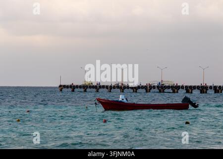 Kleines rotes Fischerboot schwimmt auf türkisfarbenem Meer in der Nähe eines langen Piers mit schwarzen Gummipuffern unter einem weichen bewölkten Himmel. Stockfoto