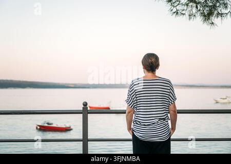 Frau, die während der frühen Morgen-Dämmerung am ruhigen Meeresufer steht. Konzept von friedlicher Achtsamkeit, Meeresflucht und stillem Reise-Moment Stockfoto