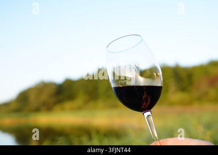 Genießt Rotwein am Fluss während des Sonnenuntergangs mit klarem Himmel und Bäumen im Hintergrund. Die Person hält ein Glas Rotwein nahe dem Flussufer. Die Szene Stockfoto