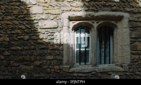 Eine Nahaufnahme fängt eine alte Steinmauer ein, die ein doppelt gewölbtes Fenster mit Schutzgittern zeigt, das in gesprenkeltem Sonnenlicht badet. Dieses Bild weckt in mir Stockfoto