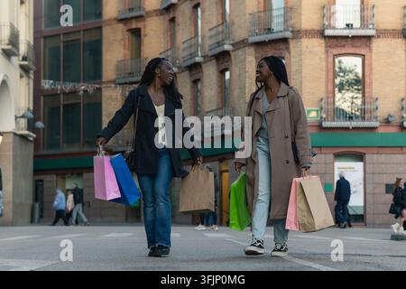 Zwei fröhliche afrikanische Frauen, die Einkaufstüten halten, gehen die Straße in der Stadt entlang Stockfoto