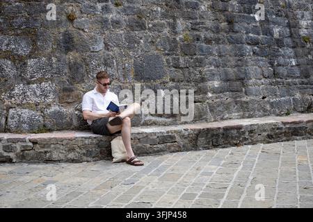 3. Juli 2026: Budva, Montenegro, junger Mann in lässiger Sommerkleidung sitzt auf einer niedrigen Steinbank und liest ein Buch, während helles natürliches Sonnenlicht t Stockfoto