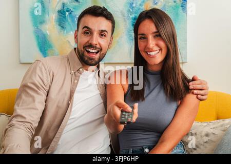 Ein fröhliches Paar sitzt zusammen auf einer Couch, lächelt in Richtung Kamera, während eine Person eine Fernbedienung hält. Die Szene vermittelt gemeinsame Unterhaltung Stockfoto
