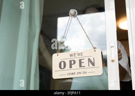Schild mit der Aufschrift „Wir haben geöffnet“ hängt an der Glastür. Eine Hand hält die Tür leicht angelehnt. Warmes Licht im Inneren lädt Kunden ein. Ideal für Geschäft, Laden oder Café pr Stockfoto