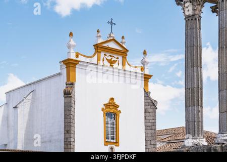 Ansicht der historischen Kirchenfassade mit gelbem Rahmen und einem Kreuz, gelegen neben den antiken römischen Säulen des Templo Romano de Evora in Evora, Ev Stockfoto