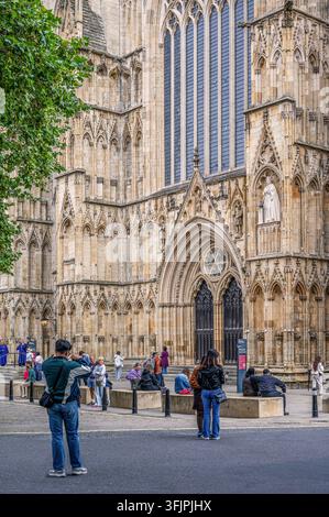 Außerhalb der York Minster und ihrer kunstvollen gotischen Architektur  Eine Menschenmenge von Touristen vermischt sich auf dem Vorplatz, sitzend und stehend. Das Minster-Personal ist an der Stockfoto