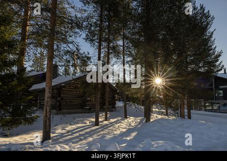 Blick auf die Levi-Blockhütte, die in einem verschneiten Kiefernwald liegt, mit einem hellen Sonnenstrahl, der durch die Bäume in Kittila, Lappi, Finnland fällt Stockfoto