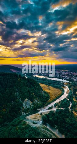 Düsterer Himmel wogt über einem Berghang und einem Fluss, der sich in einer fernen Stadt im Zwielicht schlängelt. Die Drohnenperspektive fängt kühne Farben und Tiefe ein, während Stockfoto
