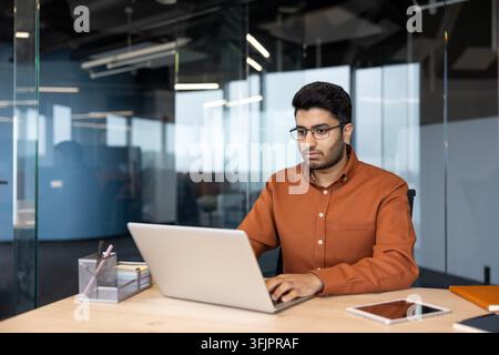 Konzentrierter Geschäftsmann arbeitet an einem Laptop in einem modernen Büro, professioneller Mann nutzt den Computer für Finanzanalysen und digitale Kommunikation an seinem Schreibtisch im Unternehmensarbeitsplatz für tägliche Geschäftstätigkeiten. Stockfoto