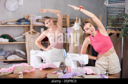 Zwei Schneiderinnen stehen in der Werkstatt und machen nach langer Arbeit eine Aufwärmübung. Näherinnen dehnen ihre Muskeln. Team von Schneidern während einer Pause bei der Ar Stockfoto