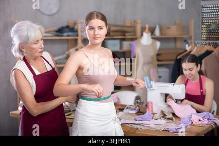Ältere Schneiderin steht neben einer jungen Frau und nimmt Maß für die Anfertigung von Kleidung vor dem Hintergrund einer Schneiderin. Handwerkerin lehrt Stockfoto