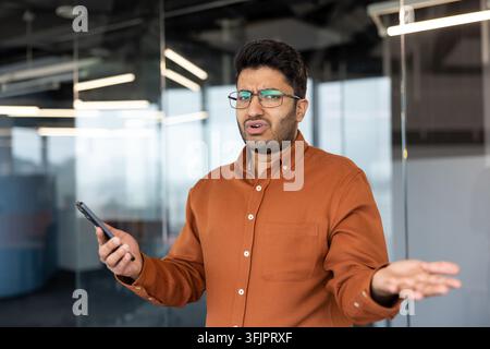 Frustrierter Geschäftsmann hält ein Smartphone in einem modernen Büro, verwirrter Berufstätiger mit Brille erlebt technische Probleme oder schlechte Nachrichten während eines Arbeitstages am Schreibtisch. Stockfoto