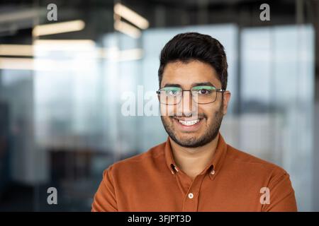 Porträt eines selbstbewussten indischen Geschäftsmannes, der in einem modernen Büro lächelt, professioneller Angestellter mit Brille und orangefarbenem Hemd für Führungspositionen und Karrierenerfolg in einem Geschäftsumfeld. Stockfoto