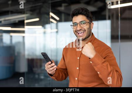 Erfolgreicher Geschäftsmann feiert Erfolg mit Smartphone im Büro, glücklicher junger Mann schaut mit Begeisterung und Siegesgeste für Geschäftswachstum auf den Handybildschirm. Stockfoto