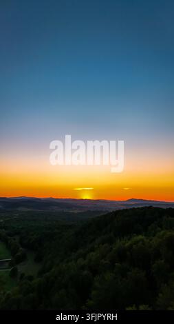 Hoch über einem dunklen Wald brennt der Horizont mit warmem Nachglühen, während sich der Himmel von Orange zu tiefem Blau verfärbt und ferne Hügel in Stille ruhen Stockfoto