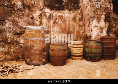 Eine Reihe alter, rustikaler Holzfässer, die an einer strukturierten Stein- und Ziegelwand in einem Keller lehnen. Ein aufgerolltes Naturfaserseil liegt auf dem Boden in Stockfoto