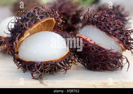 Nahaufnahme von mehreren geöffneten Rambutan-Früchten, die auf einer rustikalen Holzoberfläche angeordnet sind. Das Bild betont den Kontrast zwischen der stacheligen rotbraunen Schale und Stockfoto