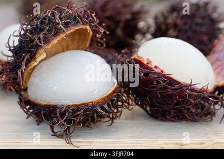 Nahaufnahme von mehreren geöffneten Rambutan-Früchten, die auf einer rustikalen Holzoberfläche angeordnet sind. Das Bild betont den Kontrast zwischen der stacheligen rotbraunen Schale und Stockfoto