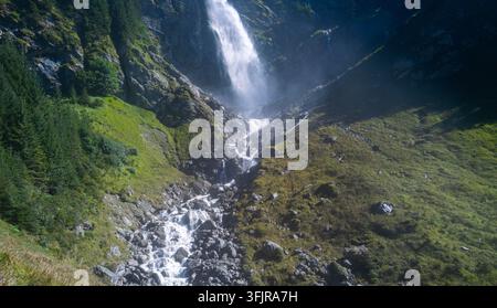 Wasserfall speist einen schnellen Bergbach. Der Wasserfall stürzt über schroffe Bergfelsen. Klares Wasser unterhalb des Wasserfalls fließt bergab. Der steinige Bach fließt Stockfoto