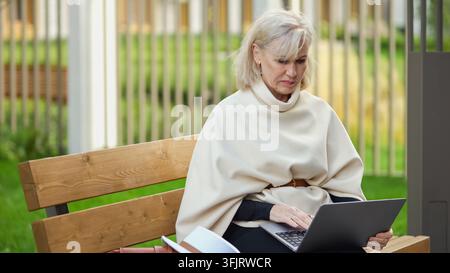 Eine Schriftstellerin tippt auf einem Laptop, während sie auf einer Bank in der Nähe von Grünzeug sitzt. Die nachdenkliche Frau hält ein offenes Notizbuch in der Nähe und bleibt konzentriert aufmerksam Stockfoto