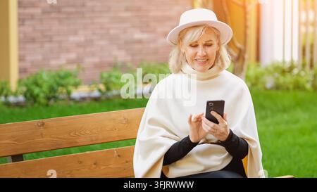 Lächelnde Seniorin bedient Smartphone neben Gartengrün. Selbstständiger Telefonnutzer hält das Mobiltelefon sicher und berührt seinen Bildschirm, präsentiert Stockfoto