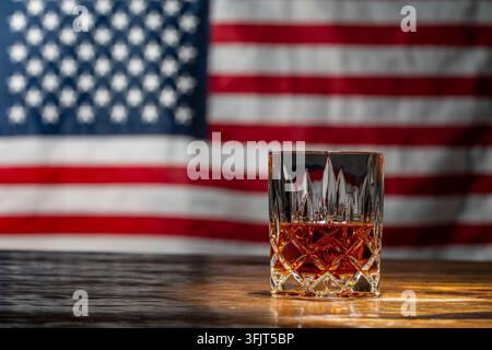 Amerikanisches Bourbon-Whisky-Glas vor Hintergrund der US-Flagge, Nahaufnahme. Kristallglas gefüllt mit bernsteinfarbenem Bourbon-Whisky steht auf Holztablett mit Unschärfe Stockfoto