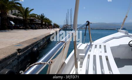 POV-Nahaufnahme vom Deck eines weißen Holz-Vergnügungsbootes, das entlang einer sonnigen Resort-Promenade mit Palmen und Café-Terrassen festgemacht ist. Ideal für den Sommer v Stockfoto