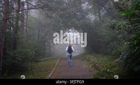 Eine Frau mit blonden Haaren geht von der Kamera weg auf einem Pfad durch einen dichten Wald. Sie trägt ein weißes langärmeliges Hemd, blaue Jeans und Stockfoto