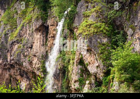 Eine natürliche Landschaft mit einem Wasserfall, der einen steilen, schroffen Felsabhang hinunterstürzt. Die Klippe besteht aus strukturiertem, braunem und grauem Stein mit tiefen Stockfoto