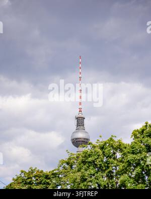 Der Berliner Fernsehturm erhebt sich über laubreiche Bäume vor einem dramatischen bewölkten Himmel im Zentrum von Berlin, Deutschland Stockfoto