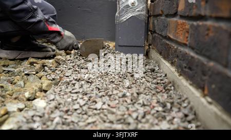 Niedriger Winkel, Nahaufnahme von Handarbeit auf der Baustelle. Eine Person in Arbeitsschutzkleidung ordnet Dränageaggregat an. Fokus auf Steine und Werkzeug mit geringer Tiefenschärfe Stockfoto