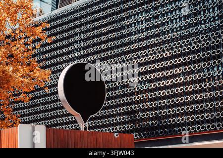Werbe-Mockup mit einer leeren schwarzen runden Werbetafel, die vor einer gemusterten Metallgebäudefassade montiert ist, umrahmt von warmem Herbstlaub für mutige Stockfoto