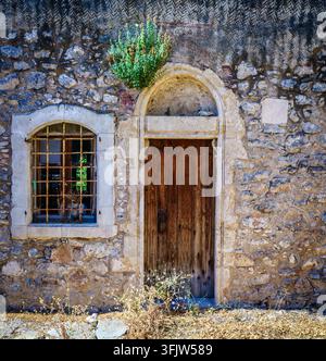 Nahaufnahme einer traditionellen kretischen Hausfassade mit gealterter Holztür, vergittertem Fenster und strukturierter Steinmauer in einem ländlichen Dorf Stockfoto