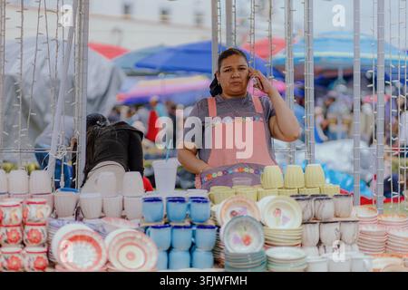 San Pedro Cholula, Mexiko - 8. September 2026: Eine Frau steht hinter einer Auslage mit Keramikbechern und -tellern auf dem traditionellen Tauschmarkt Trueque. Stockfoto