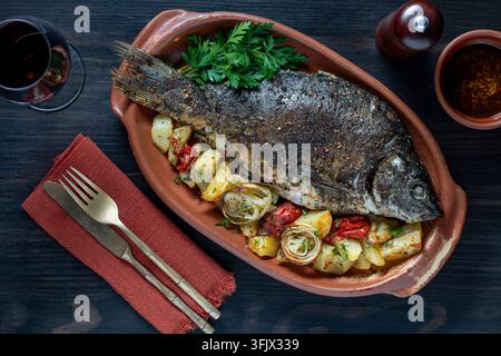 Gebackener ganzer Fisch mit Kartoffeln in Keramikschale, Ansicht von oben, Nahaufnahme. Leckerer ganz gebackener Fisch mit gebratenen Kartoffeln, Zwiebeln und Tomaten, serviert in c Stockfoto
