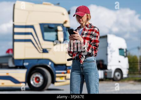 Eine Truckerin in einem karierten Hemd und Baseballmütze nutzt ihr Smartphone im Freien in der Nähe von zwei großen Sattelzügen an einem sonnigen Tag Stockfoto