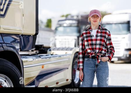 Eine selbstbewusste Frau, die ein rot-schwarz kariertes Hemd und eine rosa Baseballmütze trägt, posiert vor einer Flotte von großen Transportfahrzeugen Stockfoto