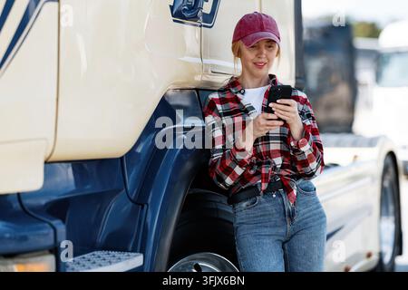 Eine Truckerin in lässiger Kleidung überprüft ihr Handy auf Nachrichten oder Navigationsanweisungen, während sie neben ihrem großen Nutzfahrzeug eine Pause einlegt Stockfoto