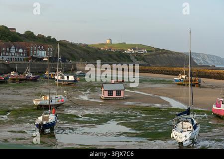 Bei Ebbe liegen Boote auf dem Schlamm im Folkestone Harbour, Kent, mit der ikonischen rosa Hütte und dem historischen Martello-Turm von East Cliff. Stockfoto