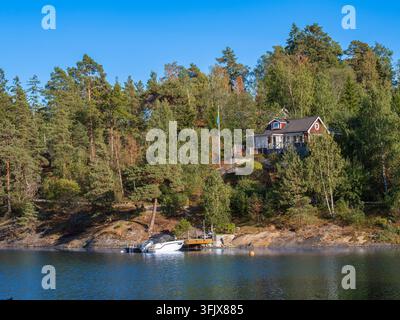 Tynningo, Schweden – 27. August 2026: Rotes Holzhaus, umgeben von Kiefern und Birken an einem felsigen Seeufer, mit einem kleinen weißen Boot, das an einem Pri Stockfoto