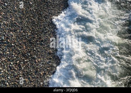 Weißer Seeschnee schwappt über eine dunkle kiesige Küstenlinie. Konzept von Meeresufer, Wasserbewegung, Wellen, natürlichen Texturen und Küstenlandschaft. Stockfoto