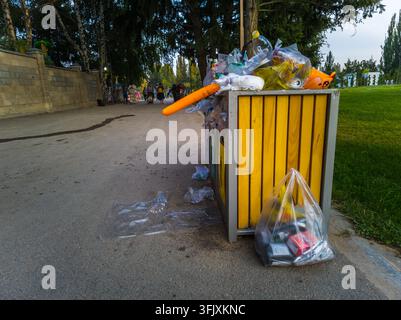 Cholpon-Ata, Kirgisistan – 26. Juli 2025: Ein gelber Abfalleimer quillt mit Müll über, umgeben von Abfall auf dem Asphaltweg. Menschen laufen in th Stockfoto