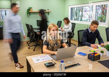 Vielfältiges Geschäftsteam, das in einem Großraumbüro zusammenarbeitet, blonde Frau konzentriert auf Laptop, umgeben von verschwommenen Geschäftsmenschen in Bewegung im Hintergrund Stockfoto