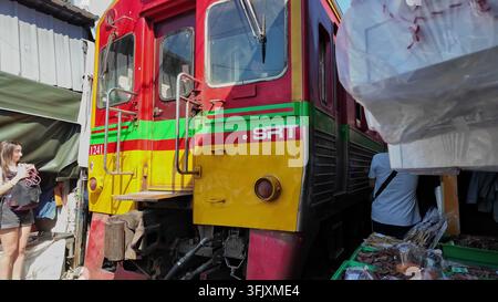 SAMUT SONGKHRAM, THAILAND - 14. MARZ 2026: Ein farbenfroher SRT-Zug fährt extrem nahe an Marktständen und Touristen am berühmten Maeklong-Eisenbahnmarkt vorbei Stockfoto