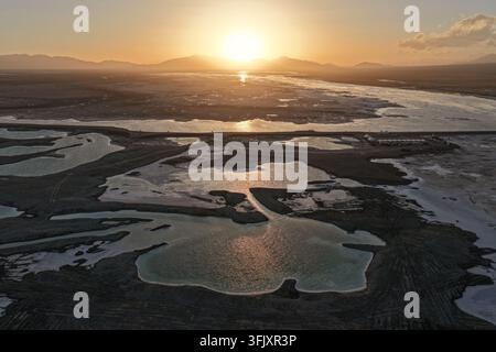Die untergehende Sonne brennt über einem Salzmarsch, das mit reflektierenden Tümpeln durchzogen ist, die das goldene Licht einfangen. Silhouettenartige Kämme umrahmen das leuchtende Hochlandbecken. Stockfoto